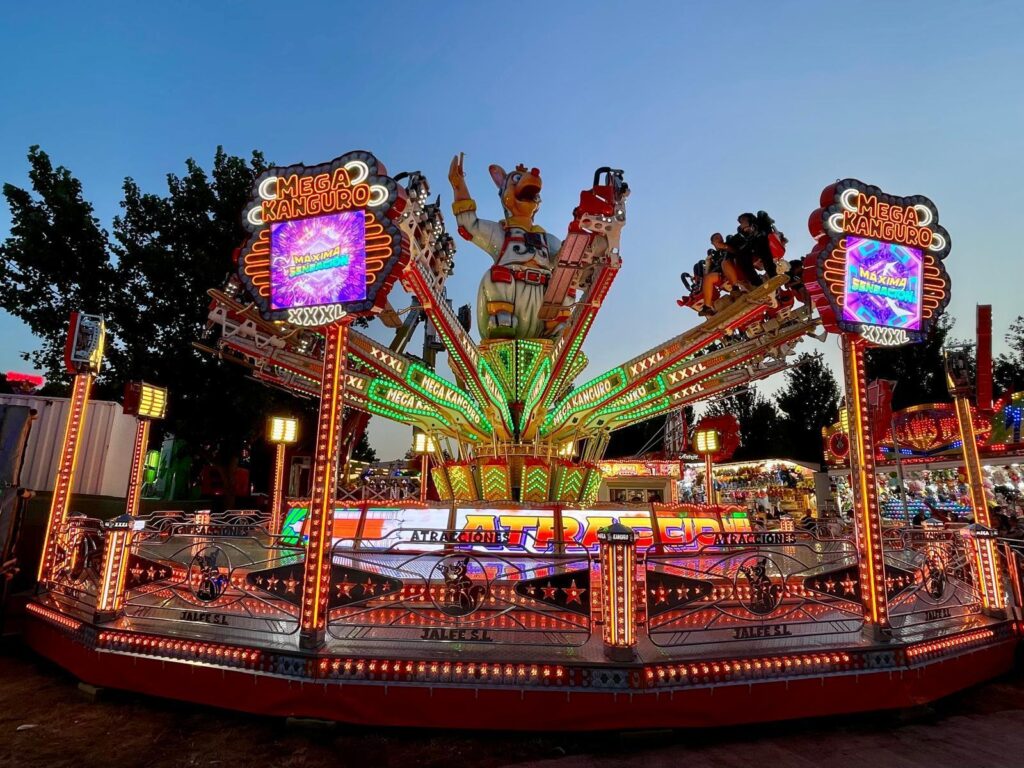 Colorful carnival ride with neon lights and large kangaroo figures at center, spinning arms and riders, fairground in the background at dusk