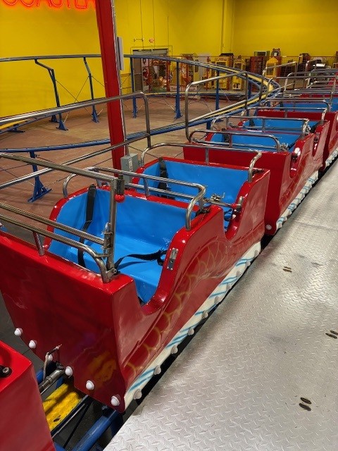 Red roller coaster cars with blue seats lined up in a station, with silver safety rails and a bright yellow wall in the background.
