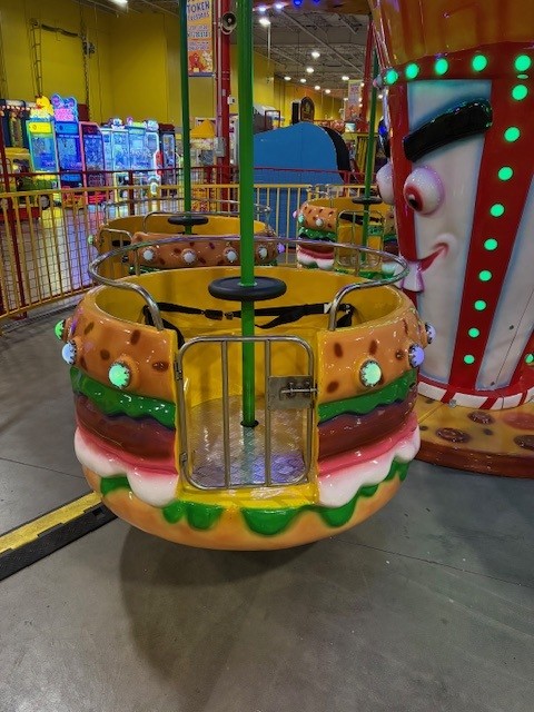 Colorful burger-themed ride car in an indoor arcade, with a green central pole and safety rails.