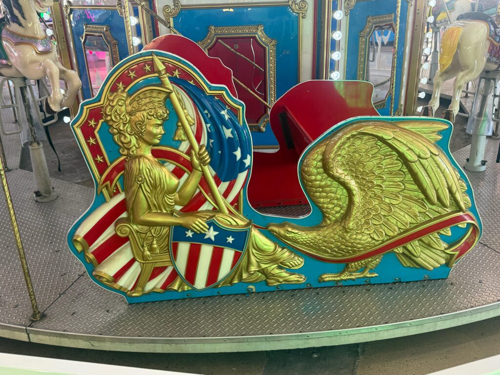 Ornate carnival ride panel with a gold eagle emblem and red, white, blue shield design on a metallic platform.