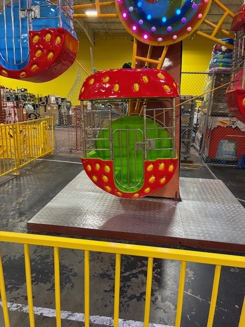Red strawberry-shaped amusement ride capsule with a green interior, mounted on a metal platform inside a colorful arcade with yellow rails.