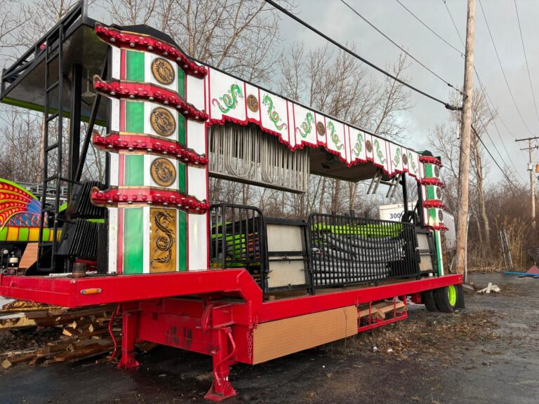 Decorated carnival ride trailer on a red platform with green-white panels and red lights, outdoors with bare trees and power lines behind.