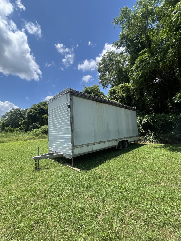 Metal cargo trailer on a grassy field beneath a blue sky with scattered clouds and trees in the background.